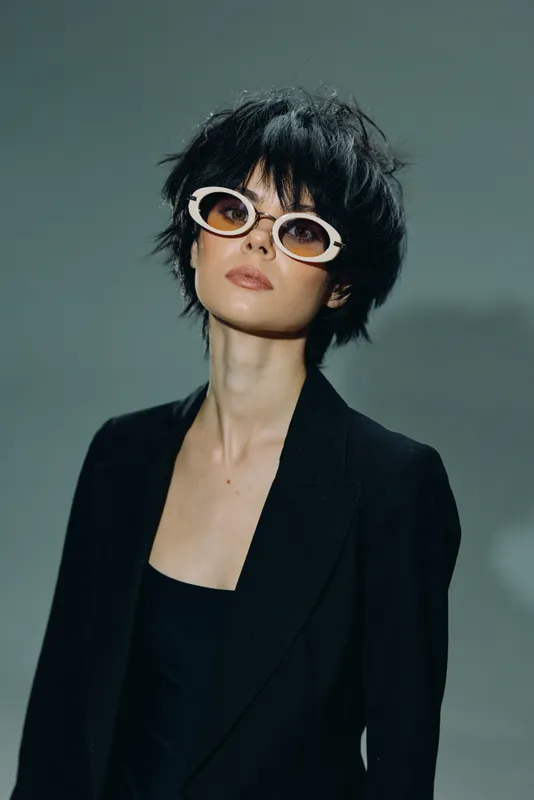 Stylish woman wearing white sunglasses and black blazer with short dark hair posing confidently indoors