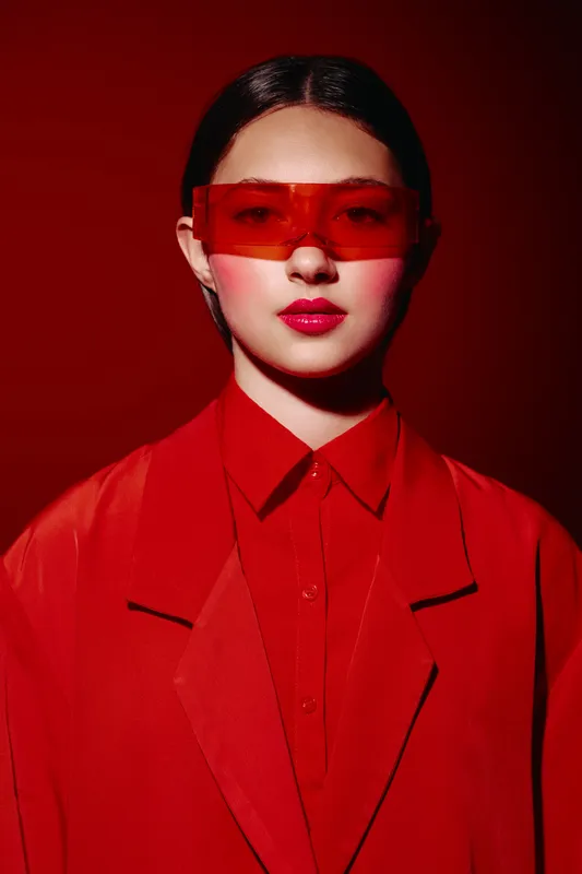 Young woman wears oversized red sunglasses and bold red lipstick in a red blazer against a red background. Creative fashion portrait focusing on beauty and style.