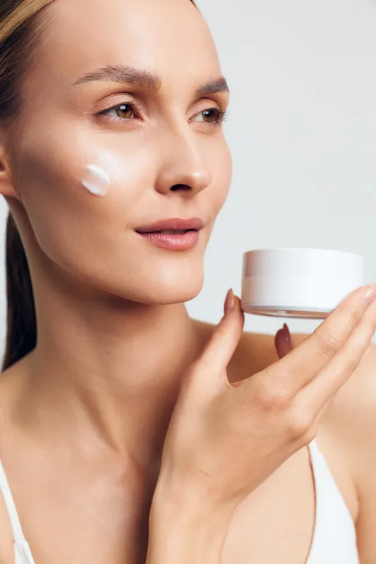 Close-up of a woman with clear skin applying white face cream on her cheek holding a jar. Concept of skincare, beauty routine, wellness, and self-care with soft natural lighting.