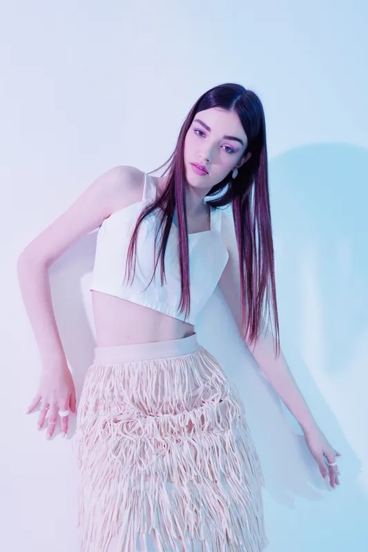 Young woman beauty fashion model posing in a white crop top and fringed skirt. Studio shot with soft pastel lighting on a plain background. Concept of style and elegance.