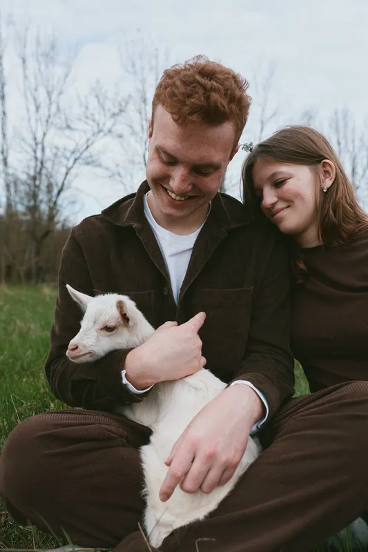 Young couple sitting outdoors with a baby goat in their arms, sharing a tender moment in casual clothing during a calm spring day.