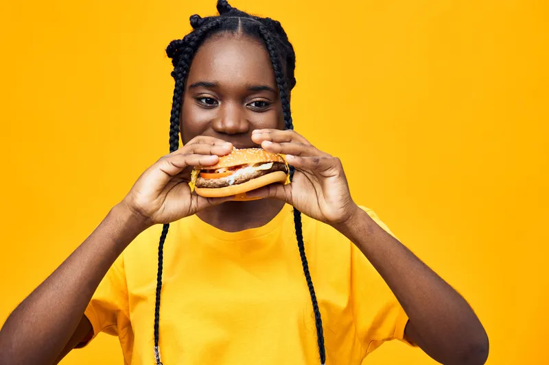 Happy young woman enjoying a delicious burger against a bright yellow background, showcasing a joyful and vibrant atmosphere related to healthy eating and indulgence