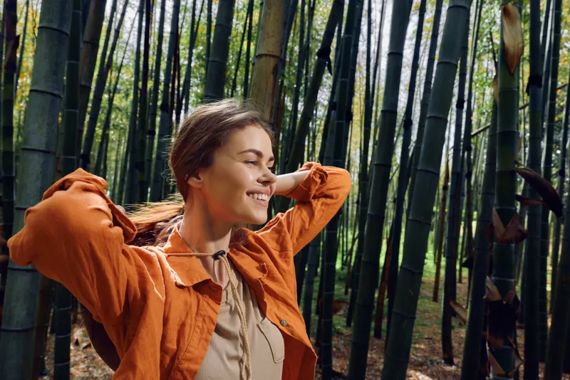 Woman in orange jacket smiling among tall bamboo forest, portrait of happy outdoor nature travel and relaxation enjoying sunlight and a peaceful green woodland scene.