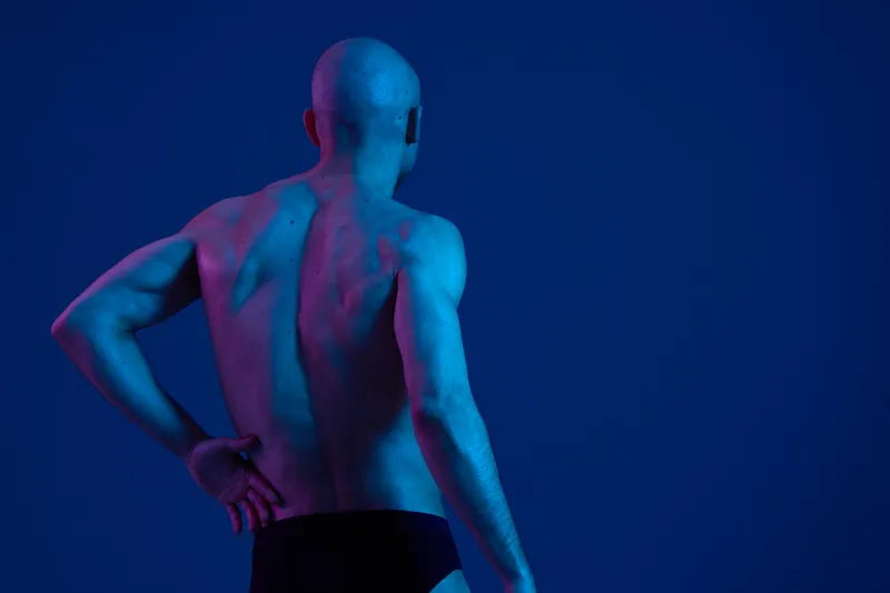 A man with a strong physique poses against a vibrant background, showcasing dramatic lighting that emphasizes his silhouette and hints at a sense of strength.