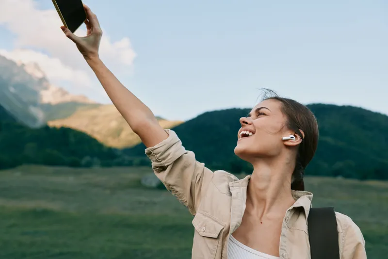 Cheerful woman outdoors, smiling into her phone, sunlight on her face, casual jacket, expansive green field, mountains in the background, relaxed lifestyle, natural mood