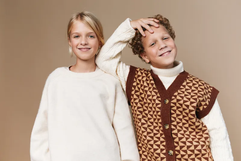Two cheerful children pose together in cozy knitwear, smiling softly against a warm beige backdrop, highlighting friendship, comfort, and playful fashion in soft earthy tones.