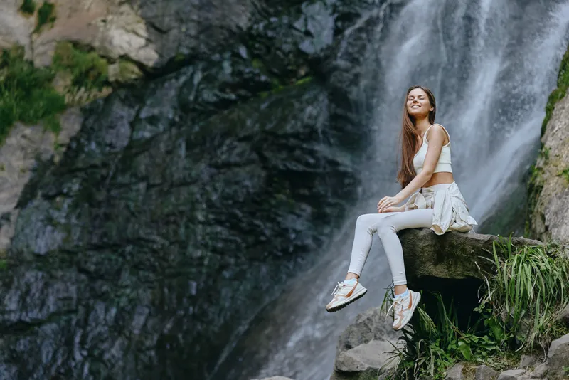 young woman sitting near waterfall in white casual clothes with relaxed smile, outdoor nature scene, fresh air, rock ledge, sunny day, peaceful moment
