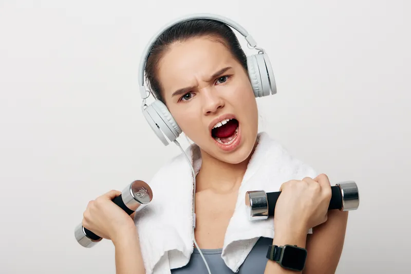 Young woman fitness portrait lifting dumbbells with towel around neck, wearing headphones and sportswatch, intense expression, studio white background, energetic workout concept