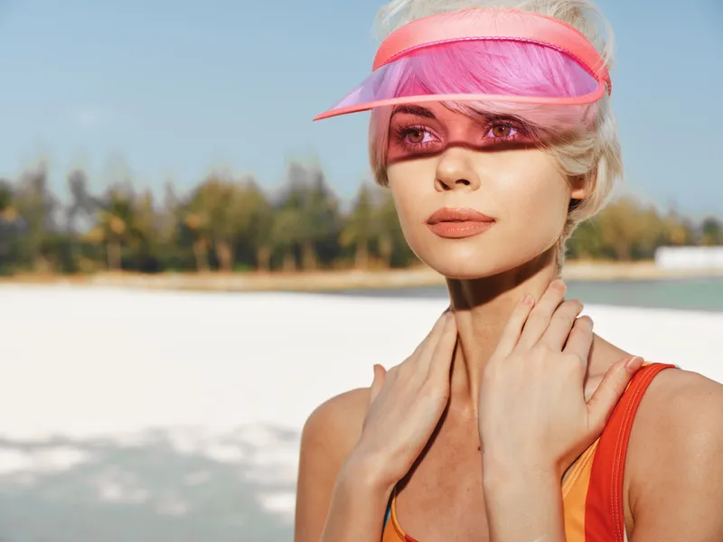 Portrait of a stylish woman wearing a pink visor on a sunlit beach, close up with smooth skin, glossy lips, and a poised gaze.
