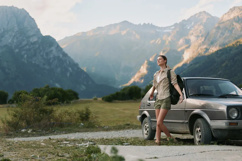 A young woman with short hair stands beside a vintage car in a rugged mountain landscape, enjoying a sunny day and a sense of freedom and exploration.