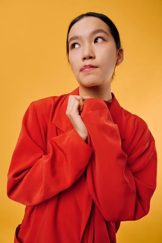Young woman in red oversized jacket posing and looking sideways on orange background. Casual fashion style with thoughtful expression and minimalistic studio setting for portrait.