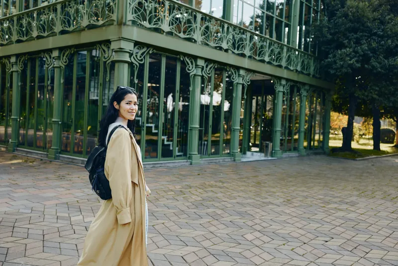 Woman in beige coat with backpack walking along a paved path beside a glass greenhouse in a park, autumn mood, candid fashion moment, outdoor travel vibe with soft daylight