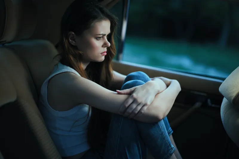 Young woman feeling sad and reflective, sitting in a car with her arms around her knees, showcasing emotions of loneliness and contemplation in a serene setting.