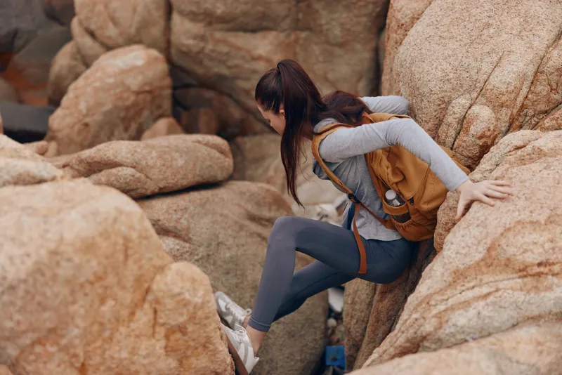 Young woman climbing rocky terrain with a backpack, showcasing determination and adventure in an outdoor setting The earthy tones highlight the natural environment