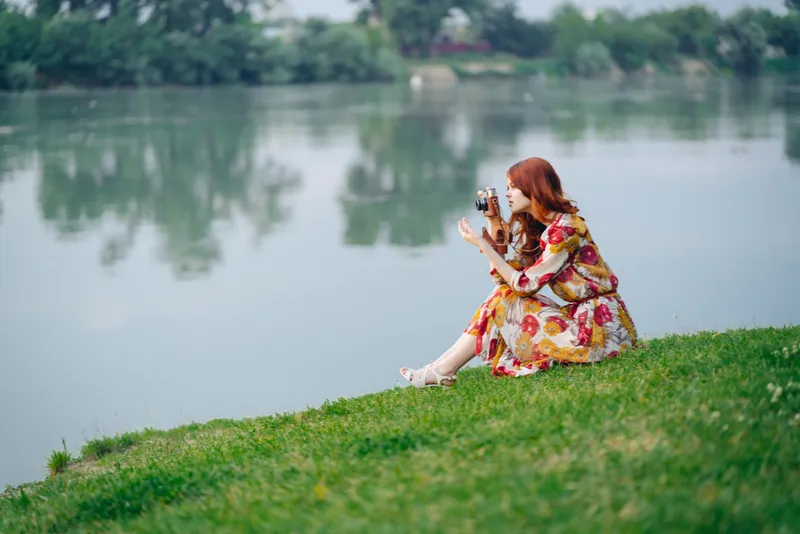 Женщина в платье с фотоаппаратом в руках стоит на берегу озера