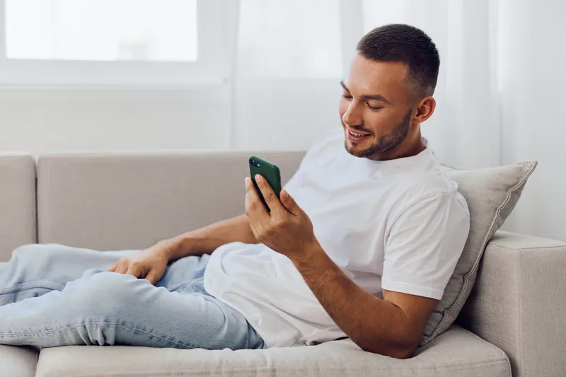 Young man relaxing on a couch, smiling while using a smartphone in a bright, cozy living room, showcasing modern lifestyle and technology trends.