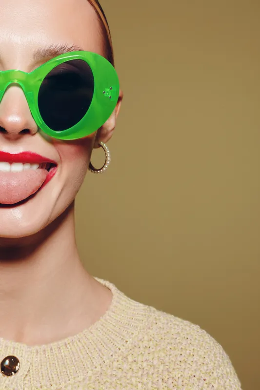 Young woman with bright green sunglasses and red lipstick sticks out her tongue playfully. Close-up portrait on beige background. Concept of fun, fashion, and summer style.