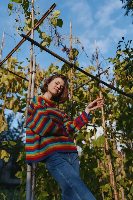 Woman colorful sweater garden vines autumn portrait, young woman in striped knitted jumper standing among trellis and foliage with sunlight and blue sky, casual jeans smile.