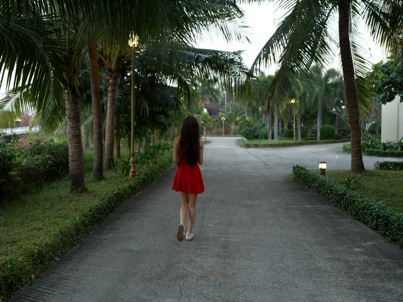 Woman walking alone on tropical pathway in red dress during dusk, surrounded by palm trees and soft evening light.
