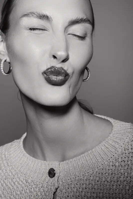 Close-up of smiling white woman with pale skin and closed eyes sending a kiss. She wears hoop earrings and knitwear, isolated on grey background. Concept of affection and beauty.
