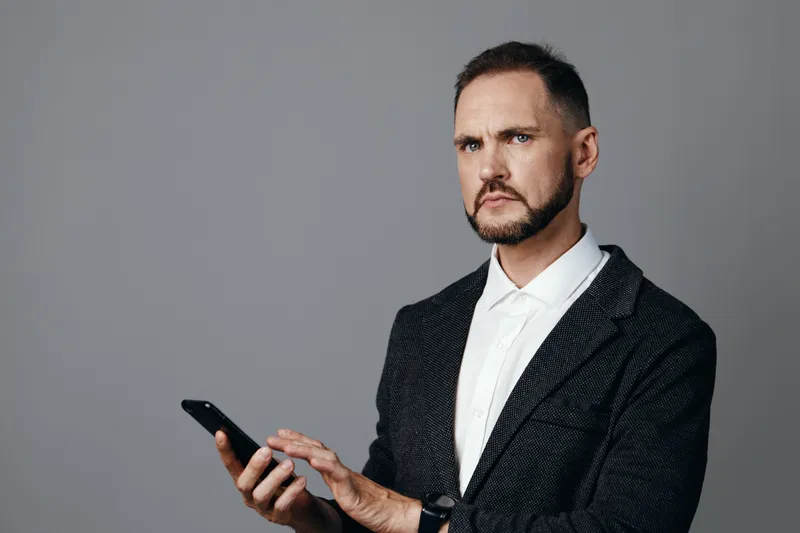 A professional businessman stands against a plain gray backdrop, holding a smartphone with a focused expression, poised for decision making and strategic communication.