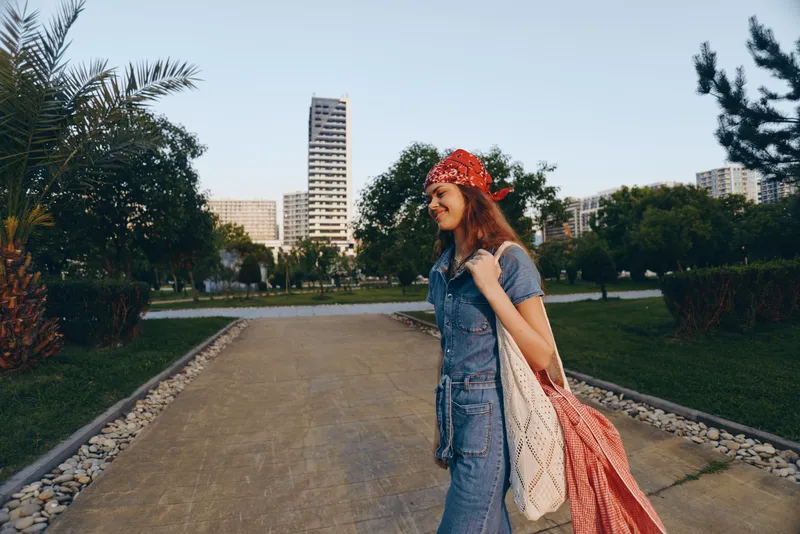 Lifestyle woman in modern boho-western denim street style walks outdoors with a red bandana and a woven bag, captured in warm film color tones.