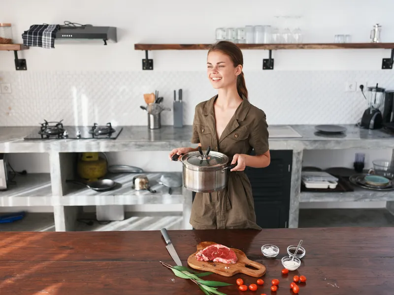 Женщина на кухне с кастрюлей в руках улыбается и готовит ужин из мяса и овощей на деревянном столе на фоне стильной кухни