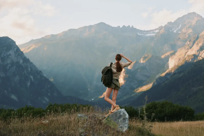 A young woman with a backpack balances on a sun-warmed rock in a wide mountain meadow, gazing toward distant peaks with a sense of exploration, freedom, and awe.