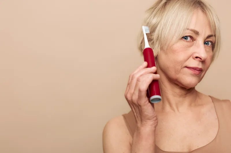 Mature woman with blonde hair holds red electric toothbrush near face on beige background. Concept of oral hygiene, dental care, personal grooming, and healthy lifestyle.