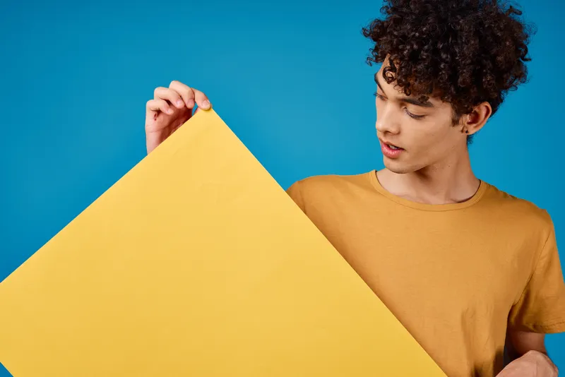 Парень с кудрявыми волосами и желтым плакатом строит гримасу на синем фоне