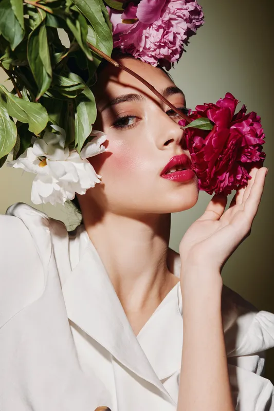 Beauty creative portrait of a woman with vibrant makeup holding red and white flowers close to her face. Fashion editoral style with dramatic lighting and elegant white clothing.