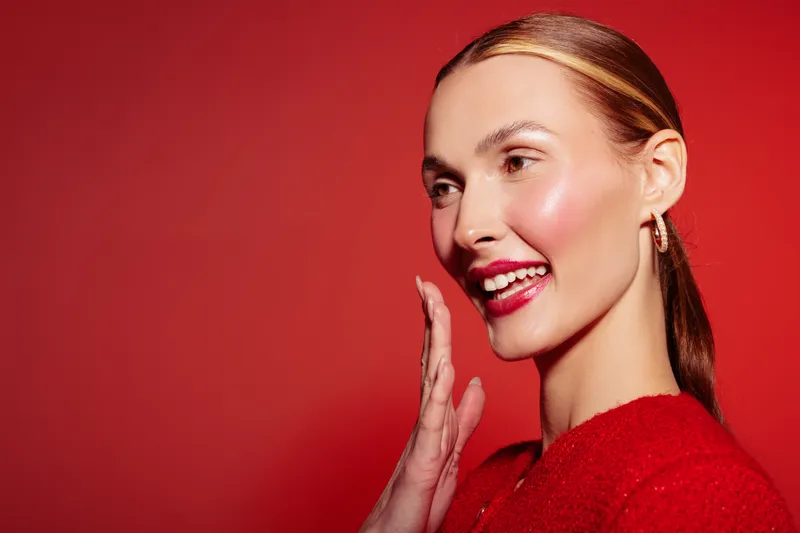 Smiling woman with shiny skin and red lipstick poses on red background. She wears red clothing and earrings, expressing happiness and confidence in a bright studio setting.