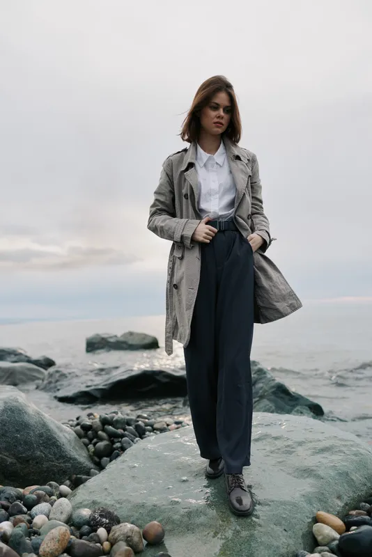 Young woman standing on rocky shore wearing long coat and formal pants. Cloudy sky and calm sea in background. Concept of fashion, confidence, and urban style outdoors.