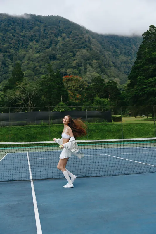 Beautiful Asian woman playing tennis on a sunny day, radiating joy and energy, surrounded by lush green nature and an inviting blue court