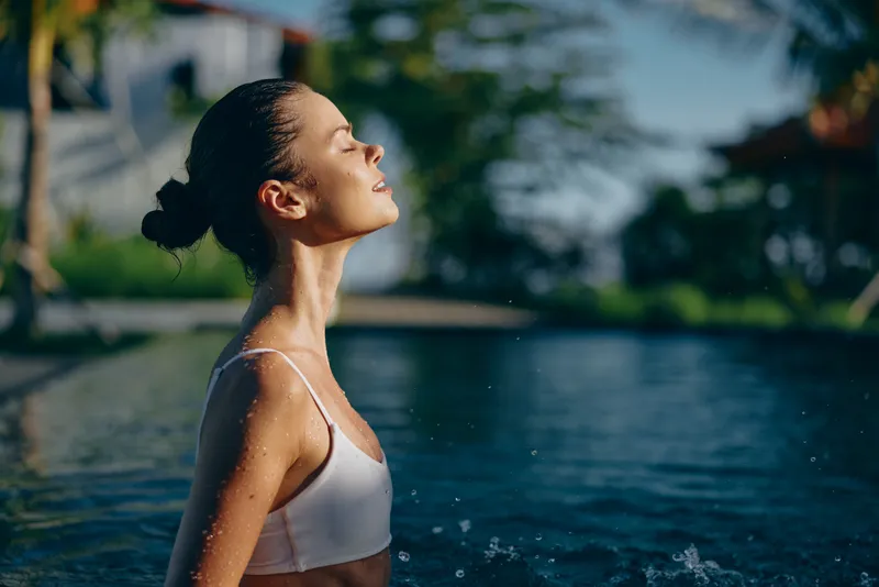 Young woman with wet hair enjoys a peaceful moment in a swimming pool with her eyes closed and face tilted towards the sunlight on a warm day.