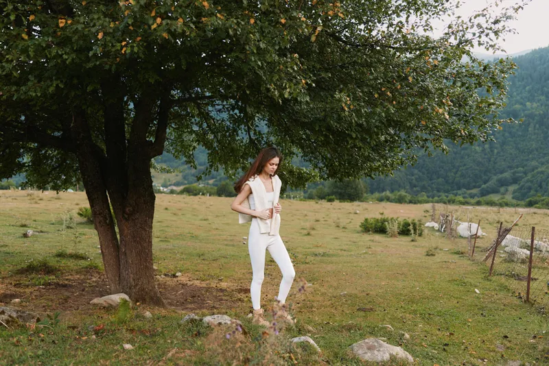 Woman standing beside a large tree in a rural meadow, wearing white clothing. Lifestyle portrait outdoors, peaceful countryside scene, natural light, relaxed pose, simple fashion.