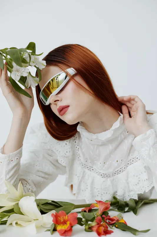 Fashionable young woman with red hair wearing stylish sunglasses, surrounded by colorful flowers, showcasing a modern, fresh look