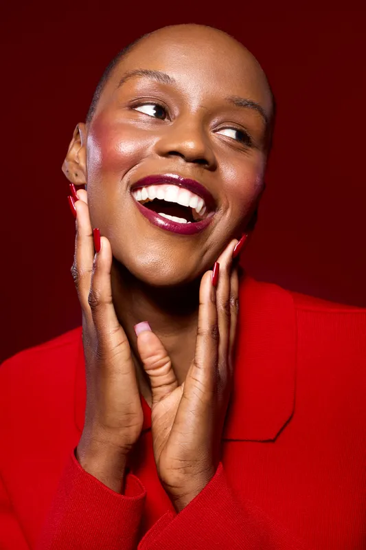 Smiling black woman with dark skin and bald head wears a red jacket, showing joyful expression and manicured nails. Studio portrait on red background with beauty and fashion concept.
