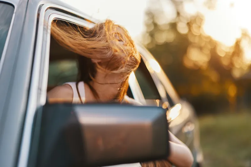 Young woman enjoying the breeze with her hair flowing out of a car window during sunset, capturing a sense of freedom and adventure in nature.