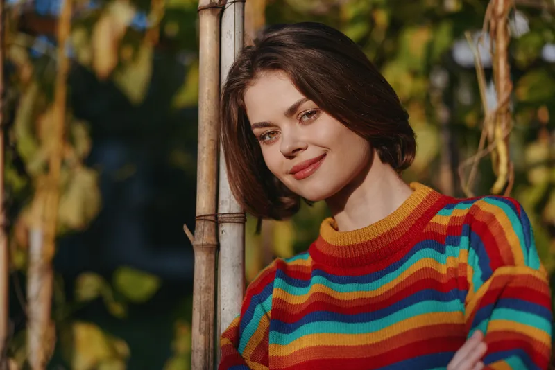 Woman portrait smile colorful sweater outdoor nature, young woman leaning on a bamboo pole in a garden with warm sunlight, cozy striped knit and relaxed confident cheerful expression.