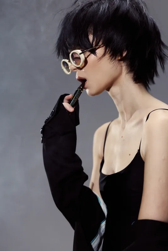 Stylish woman with short black hair wearing glasses and black outfit, thoughtful and modern look with muted background and soft lighting.