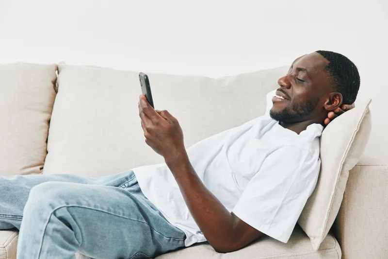 Young African man relaxing on a couch, smiling while using a smartphone in casual attire, conveying joy and comfort in a modern living room