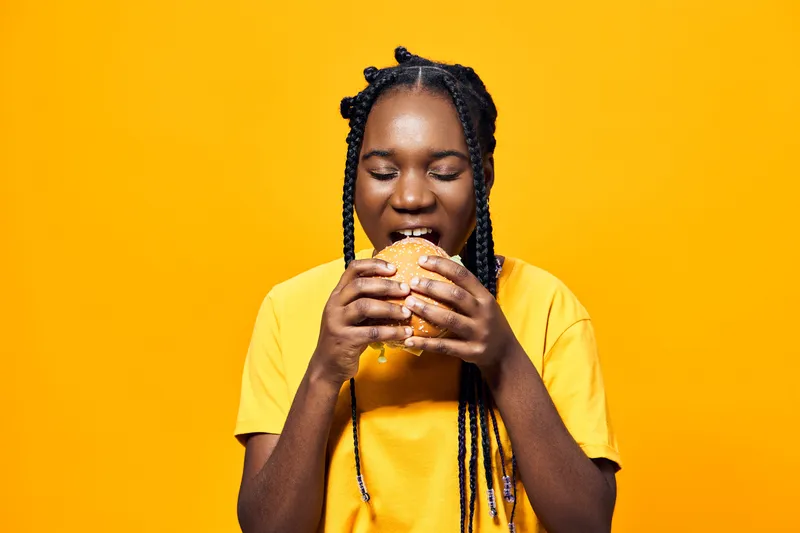 Happy young woman enjoying a delicious burger against a bright yellow background, showcasing vibrant colors and a fun, playful concept related to fast food and enjoyment
