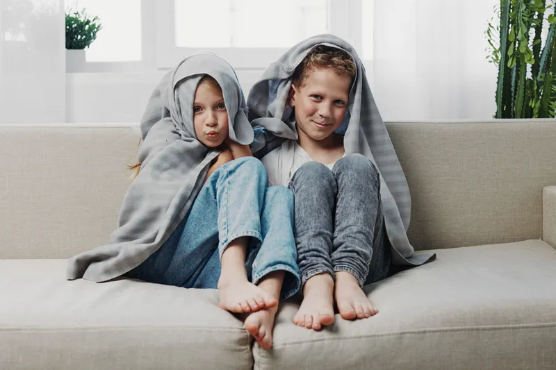 Two children sit closely on a beige sofa covered with grey blankets over their heads, barefoot and relaxed in a bright modern living room with plants. Concept of childhood, comfort, and play.
