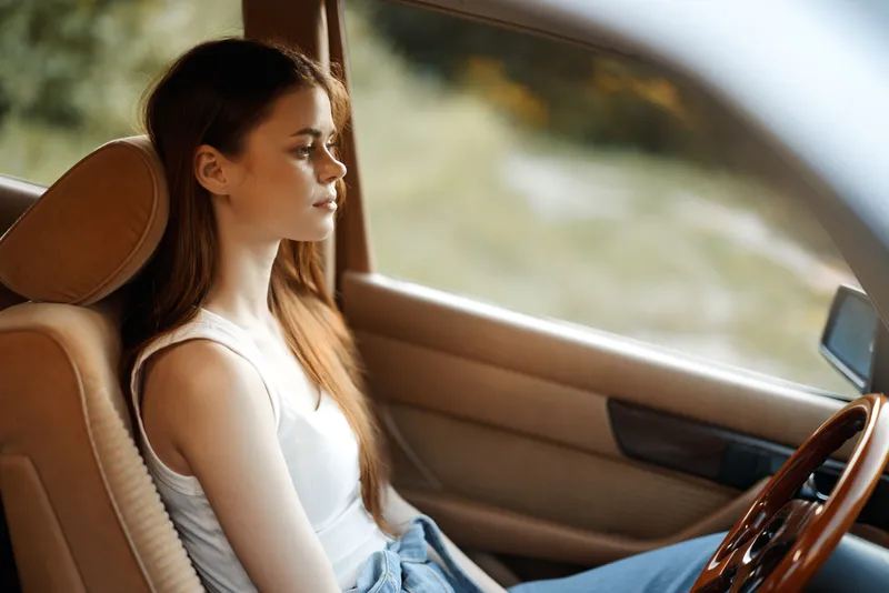A young woman with long brown hair sitting in a car, expressing calmness and contemplation, surrounded by nature with soft sunlight filtering through, reflecting a serene lifestyle.