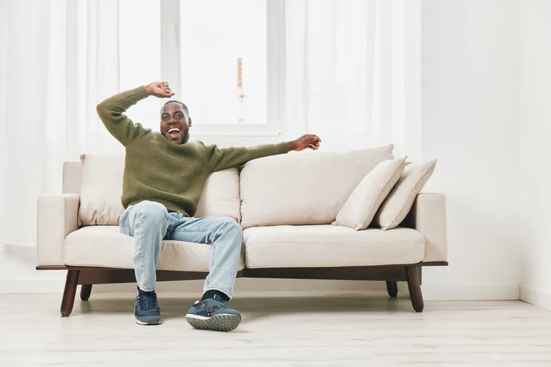 Happy young Black man sitting on a beige couch, wearing a green sweater and jeans, expressing joy and relaxation in a bright, minimalist living room