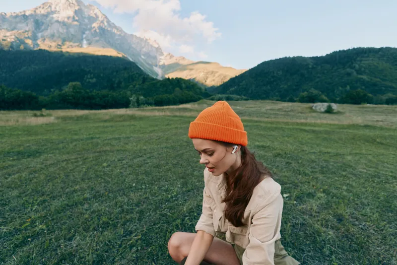 Young woman wearing orange knit hat sitting on grass in mountain landscape, casual outfit, peaceful expression, early morning light, natural scenery