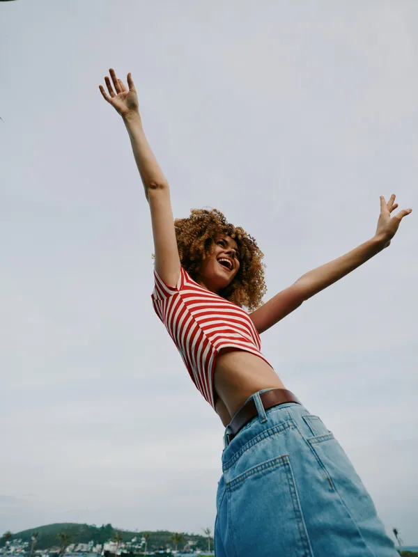 A joyful person leaps with arms raised against a bright blue sky, wearing a striped red top and denim jeans, capturing a moment of freedom, energy, and carefree summer spirit and joy.