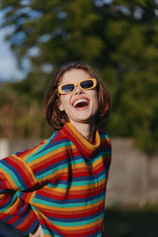 Woman smiling in yellow sunglasses and a colorful striped sweater outdoors, joyful young adult portrait laughing in casual outfit, short hair and playful pose for lifestyle fashion photography.