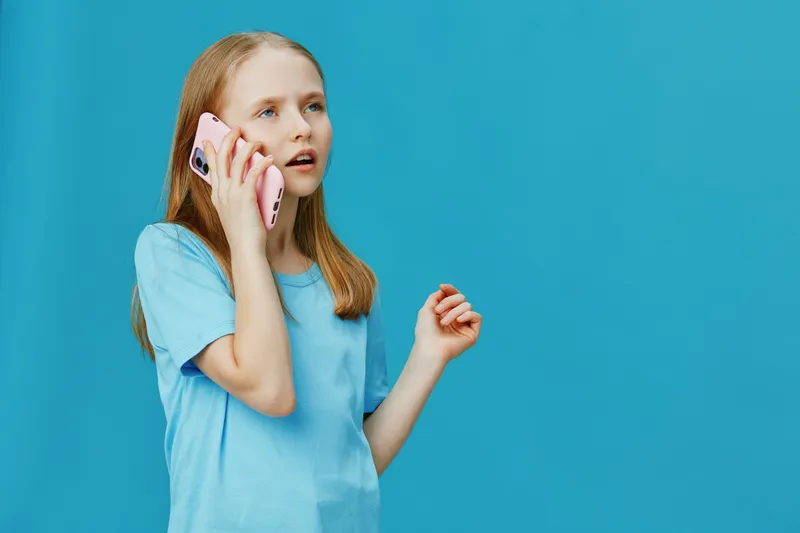Young girl in a blue shirt talking on a pink phone against a bright blue background, expressing curiosity and engagement
